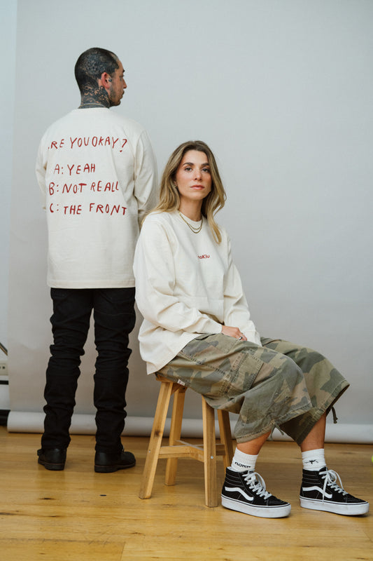 Two people, one standing one sitting down in the same light beige long sleeve shirt with red text