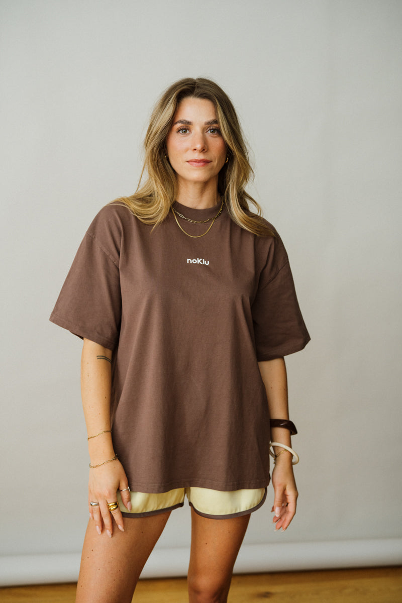 Woman wearing a brown oversized t-shirt with a visible brand logo against a plain background