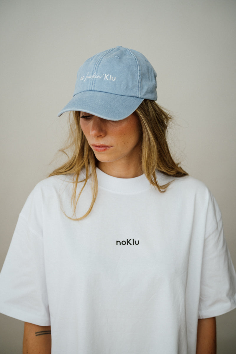 Person wearing a light blue cap and white t-shirt with 'noKlu' branding on a neutral background