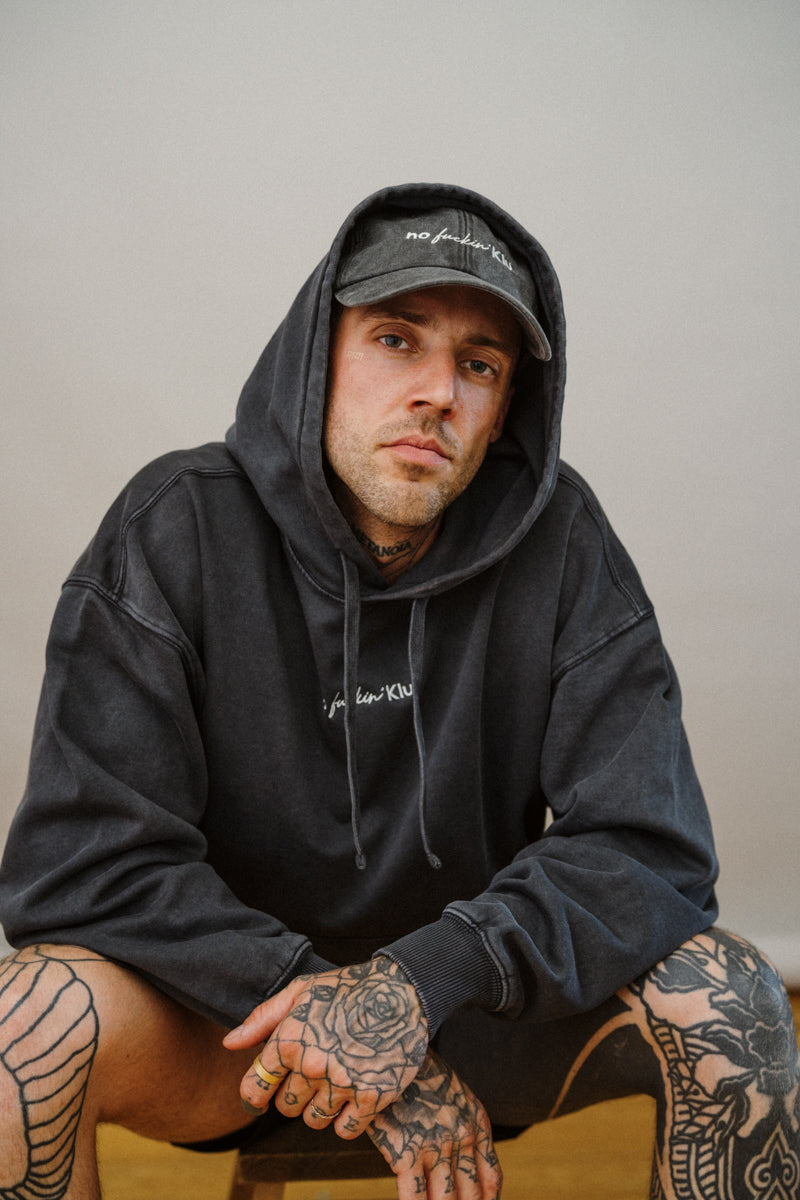 Person wearing a black hoodie and cap with tattoos on arms and legs, sitting against a plain background