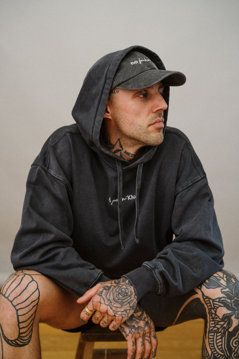 Man wearing a dark grey hoodie and cap with tattoos on his arms and legs, sitting against a plain background.