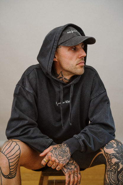 Man wearing a dark grey hoodie and cap with tattoos on his arms and legs, sitting against a plain background.