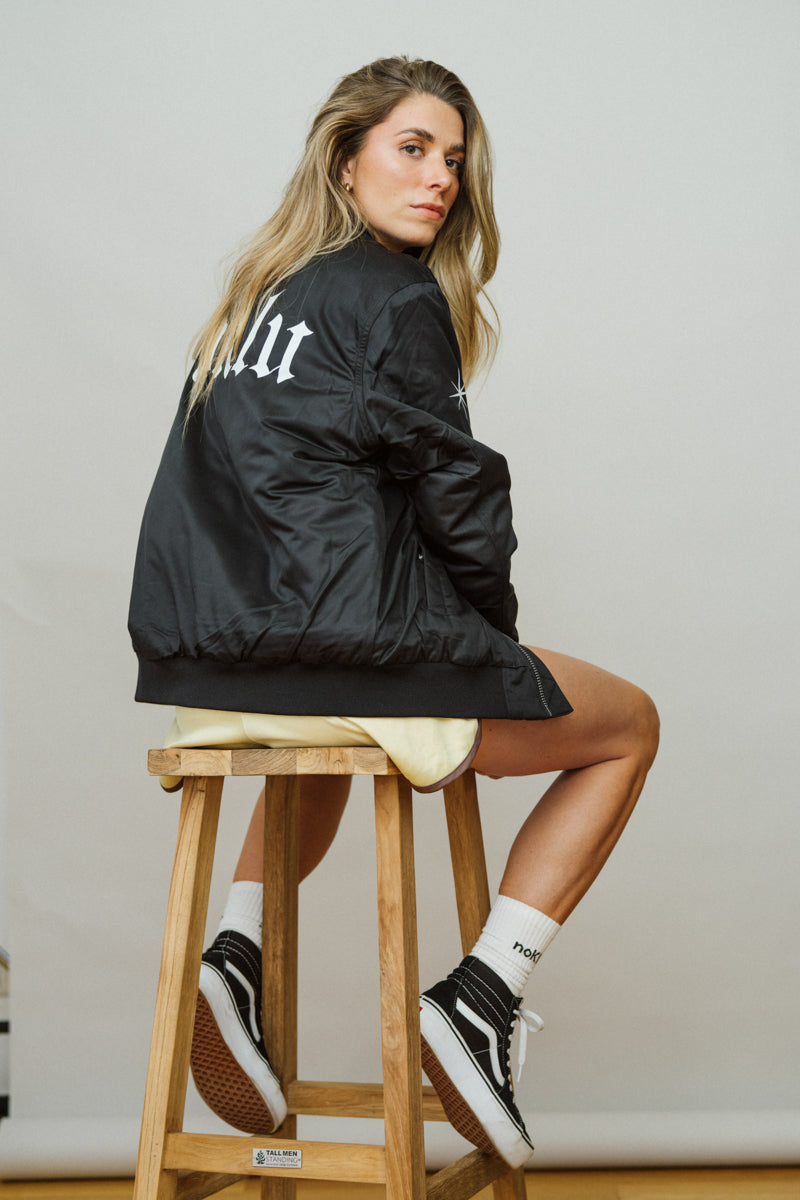 Woman sitting on a wooden stool wearing a black jacket with text, white shorts, and black sneakers.