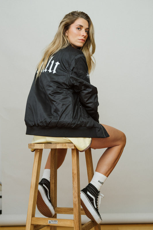 Woman sitting on a wooden stool wearing a black jacket with text, white shorts, and black sneakers.
