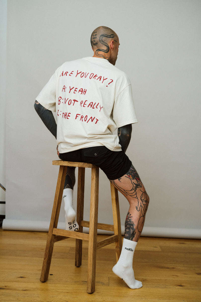 Person sitting on a wooden stool wearing a white t-shirt with text and black shorts.