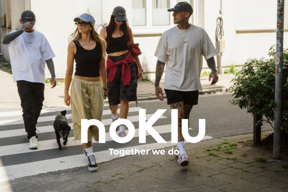 Four people walking outdoors on a city street crosswalk, wearing oversized white t-shirts, black shorts, and caps. A small black dog walks beside them.